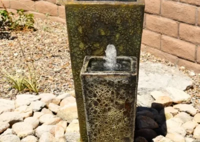 A Sculptural Twin Column Fountain By Sonoran Gardens Inc., With Bubbling Water Over Glazed Ceramic Surfaces, Nestled In River Rock For A Tranquil Desert Accent