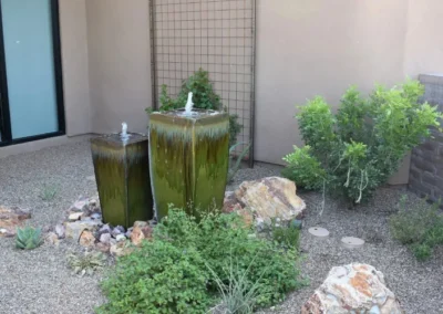 Pair Of Glazed Ceramic Water Fountains With Green Ombré Finish, Surrounded By Decorative Rock And Desert Plants Against A Stucco Wall And Trellis