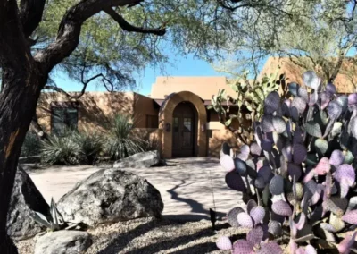 Southwestern Style Tucson Home With Arched Entry, Surrounded By Native Desert Landscaping Including Large Boulders, Purple Prickly Pear Cactus, And Mesquite Trees