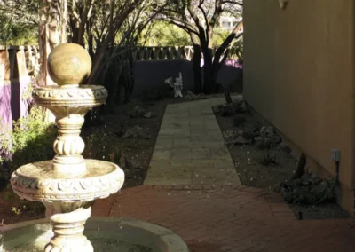 A tiered stone fountain in the foreground of a landscaped garden pathway with a small angel statue in the distance