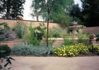 A scenic tiered fountain centerpiece in a landscaped backyard with yellow flowers and stone walls