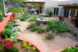 Tucson courtyard garden with colorful flowers, agave, red stucco border walls, and a tiered stone fountain at the center of a gravel landscape.